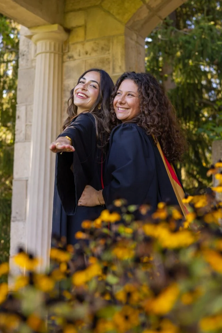 Two women in graduation gowns standing behind yellow flowers with a stone structure and trees in the background. The photo is from Guelph Grad Photos. The background is the University of Guelph Campus.