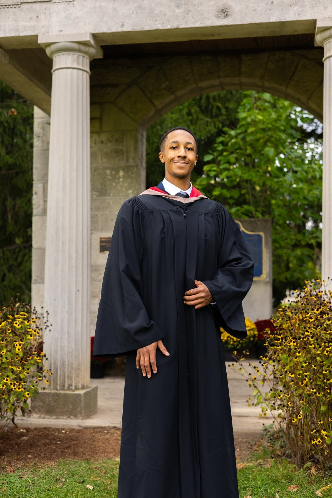 A professional photo of a young man Graduate in a cap and gown, standing outdoors under a stone archway. The photo is from Guelph Grad Photos. The background is The Portico on the University of Guelph Campus.