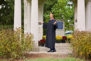 Person in graduation gown standing in front of a classical building with columns and flowers.