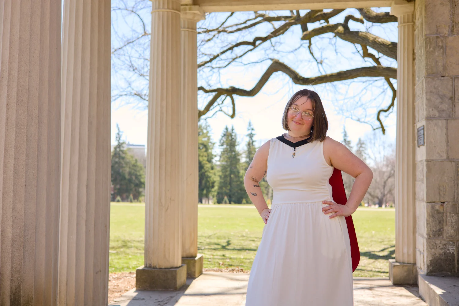 A professional photo of a Guelph grad on the University of Guelph campus, in a white dress standing in front of stone columns with a scenic background near The Portico. The photo is from Guelph Grad Photos.