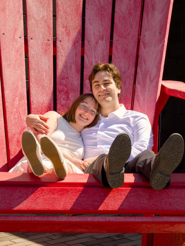 Two people sitting on a red bench with a bright background, The photo is from Guelph Grad Photos. The background is the University of Guelph Campus.