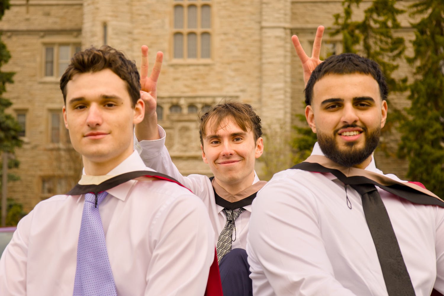 Three men in formal attire with suspenders and ties standing in front of a brick building. The photo is from Guelph Grad Photos. The background is the University of Guelph Campus.