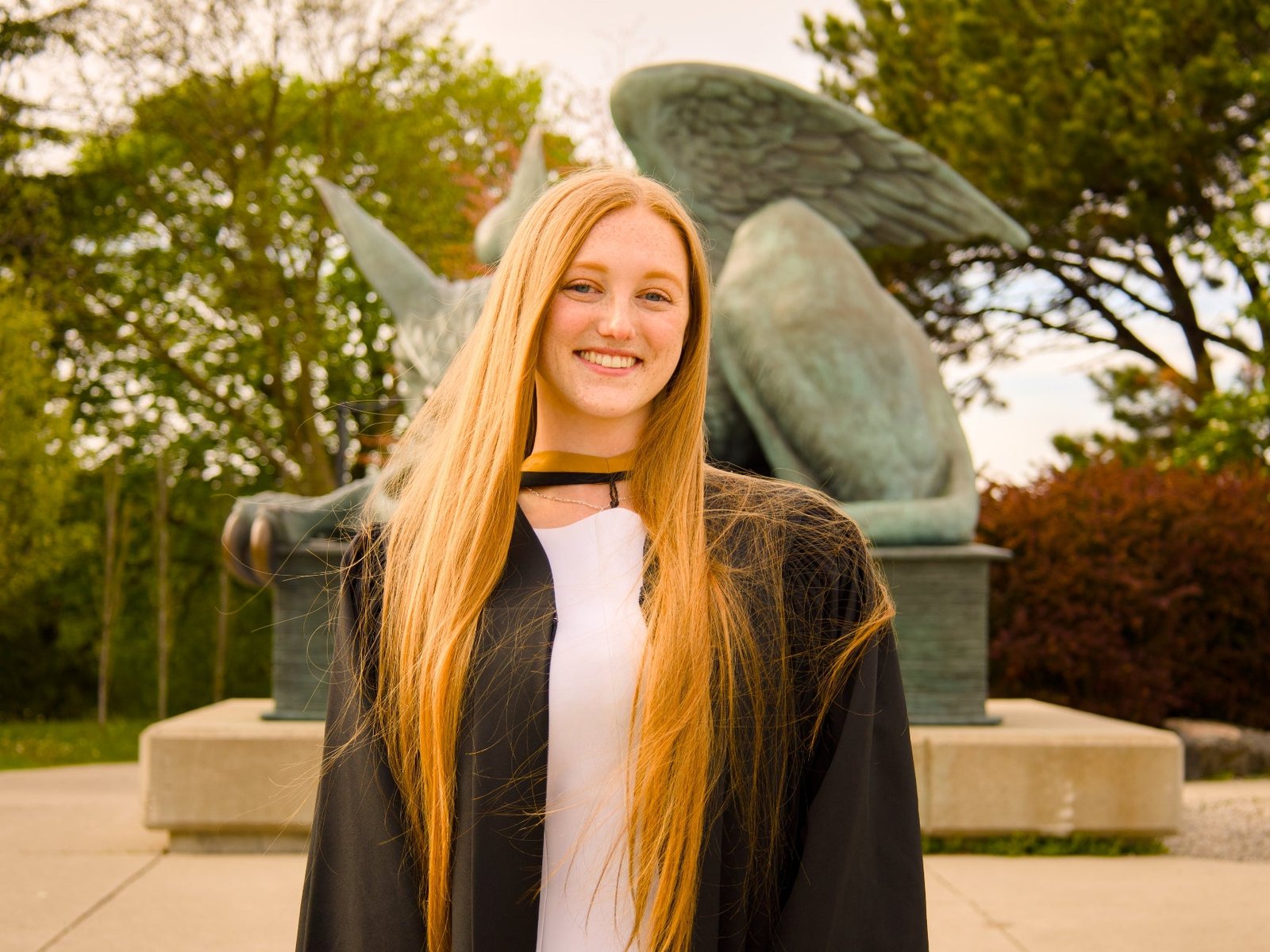 Person in graduation gown standing in front of a statue with trees in the background
