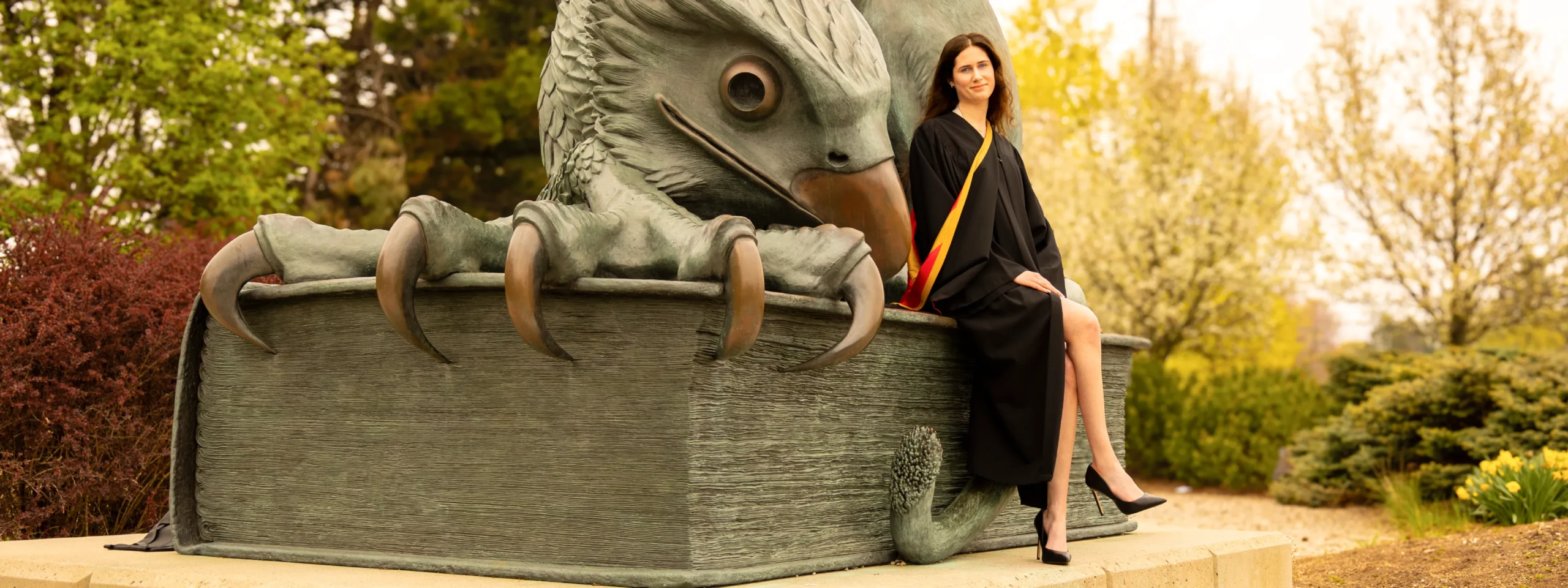A woman in a graduation gown sitting on the Guelph Gryphon sculpture on Campus