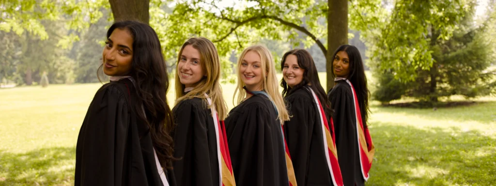 Five University of Guelph graduates in black convocation gowns and coloured hoods lined up on a sunlit campus green