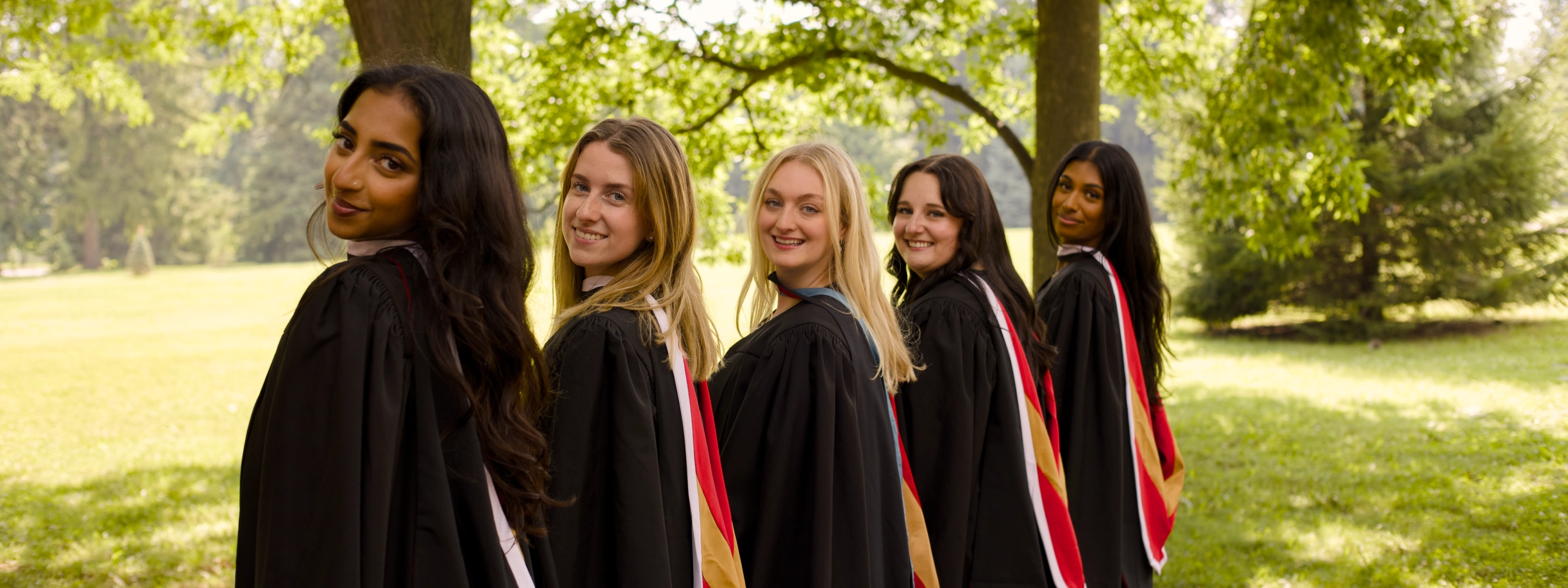 Five University of Guelph graduates in black convocation gowns and coloured hoods lined up on a sunlit campus green