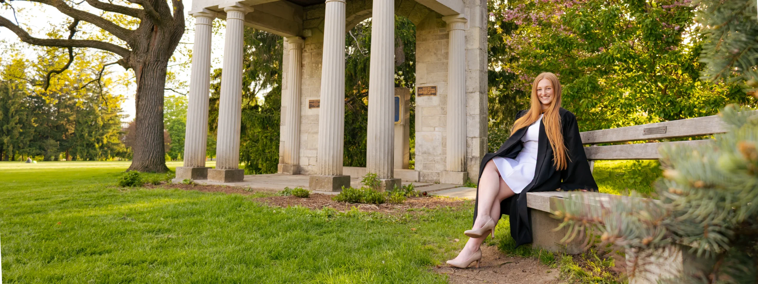 University of Guelph graduate in a black convocation gown seated by the Portico on Johnston Green in spring