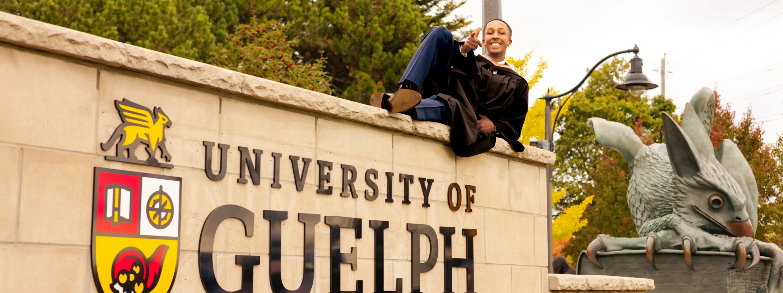 University of Guelph graduate sitting on the campus entrance sign at Gordon Street with the Gryphon statue behind him