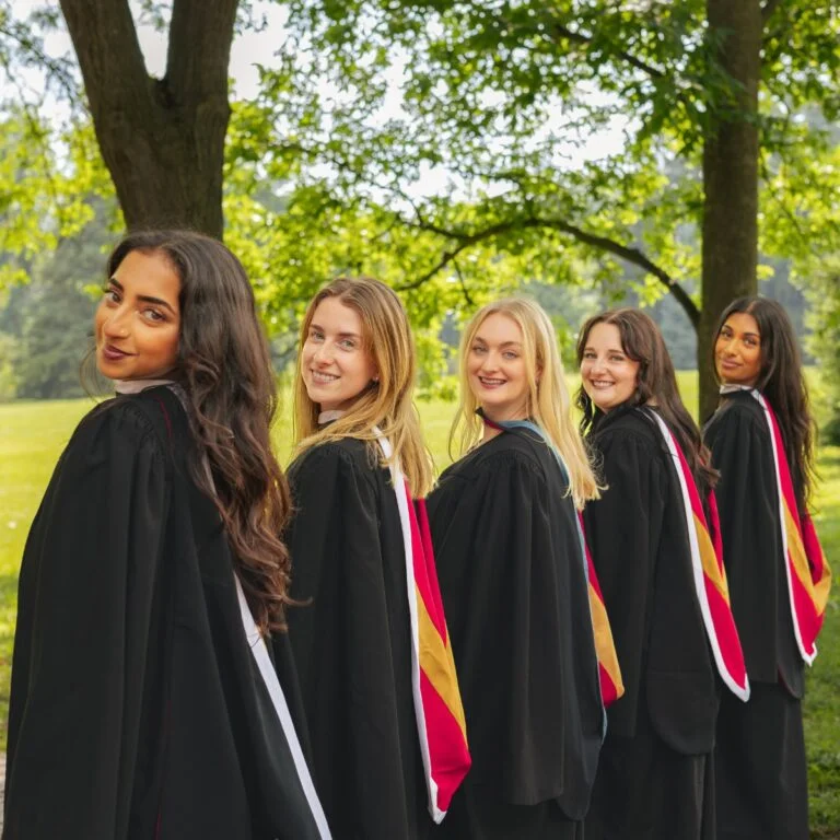 Five University of Guelph graduates in black convocation gowns and coloured hoods lined up on a sunlit campus green