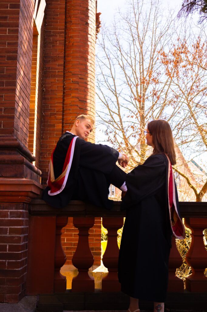 A graduate seated on a red brick balustrade holding hands with another graduate standing beside a brick column.
