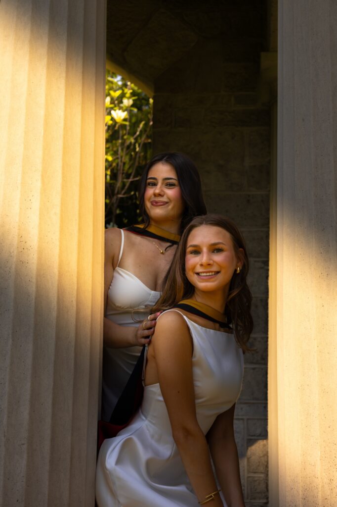 Two graduates in white dresses with black and gold hoods smiling between two stone columns in warm evening light.