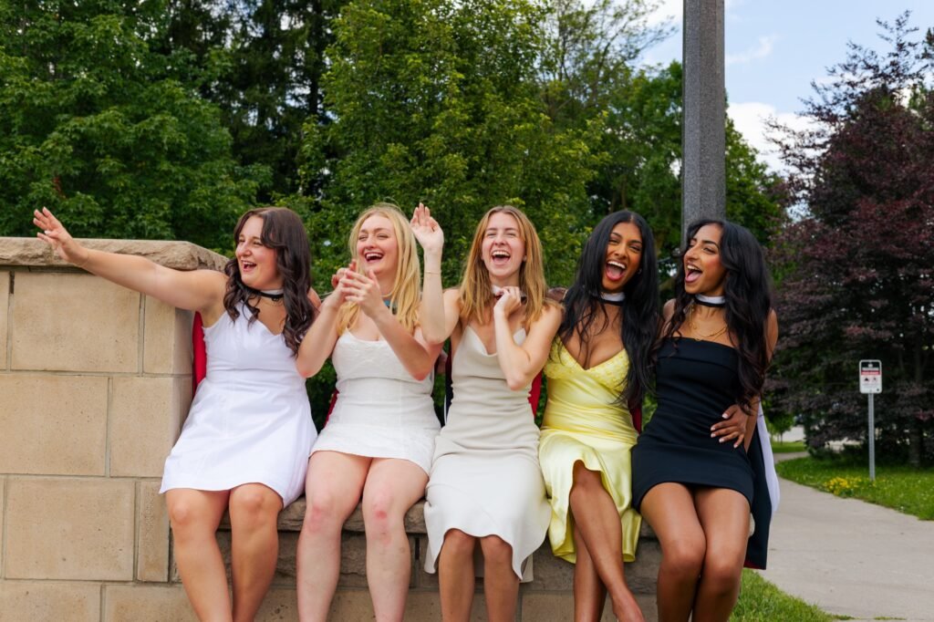 Five graduates in dresses with academic hoods draped behind them laughing together on a stone wall.