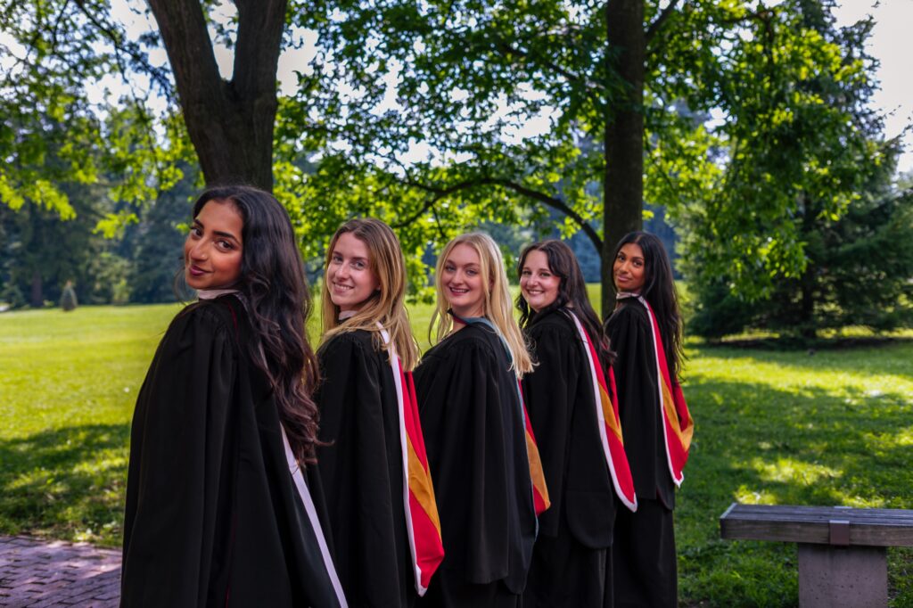 Five graduates in black gowns with red and gold hoods standing in a line and looking back over their shoulders under large trees.