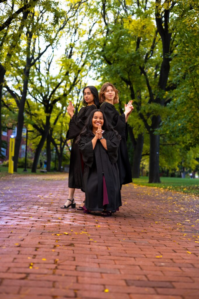 Three graduates in black gowns striking playful poses on a tree-lined brick walkway at the University of Guelph.