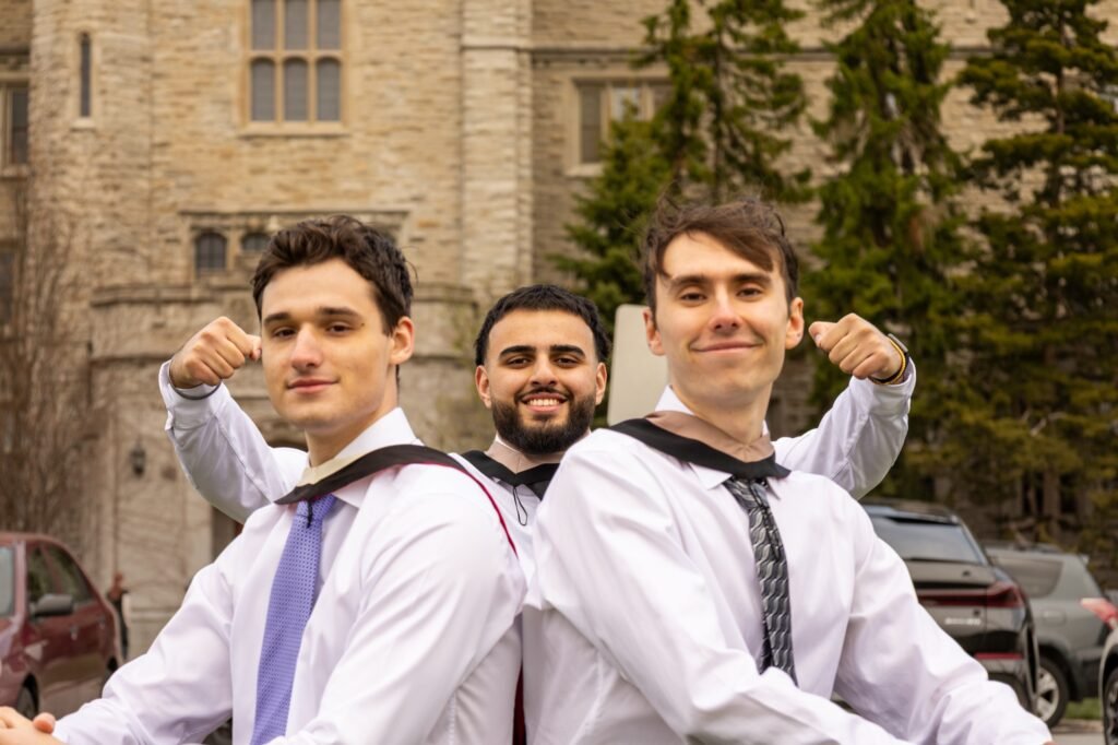 Three graduates in white shirts and academic hoods flexing their arms back to back in front of a stone building at the University of Guelph.