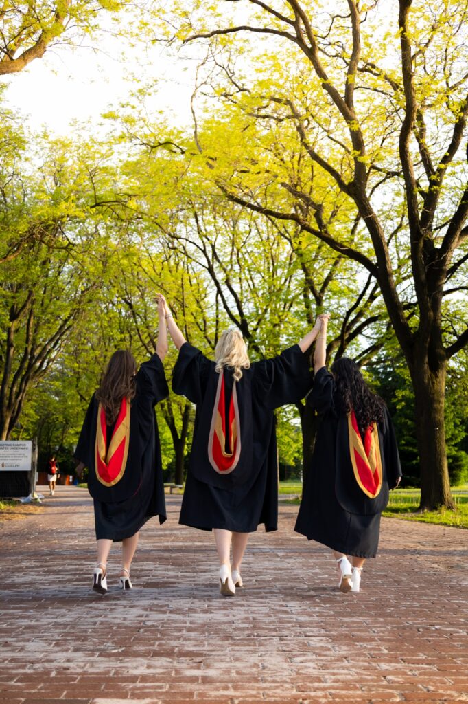 Three graduates in black gowns with red and gold hoods raising joined hands while walking away down a brick walkway at the University of Guelph.