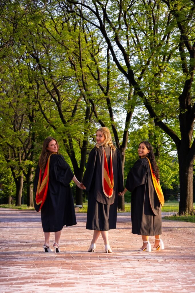 Three graduates in black gowns with red and gold hoods holding hands and looking back on a tree-lined brick walkway at the University of Guelph.