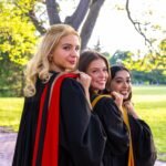 Three graduates in black gowns with red and gold hoods looking back over their shoulders on a brick path with a green lawn behind them.