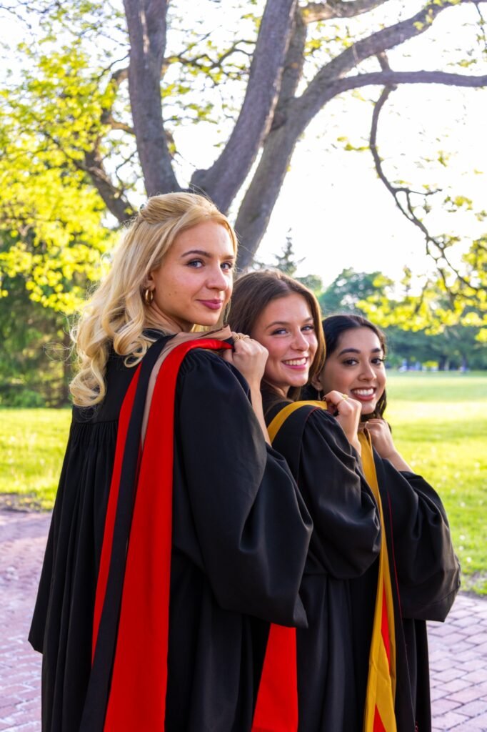 Three graduates in black gowns with red and gold hoods looking back over their shoulders on a brick path with a green lawn behind them.