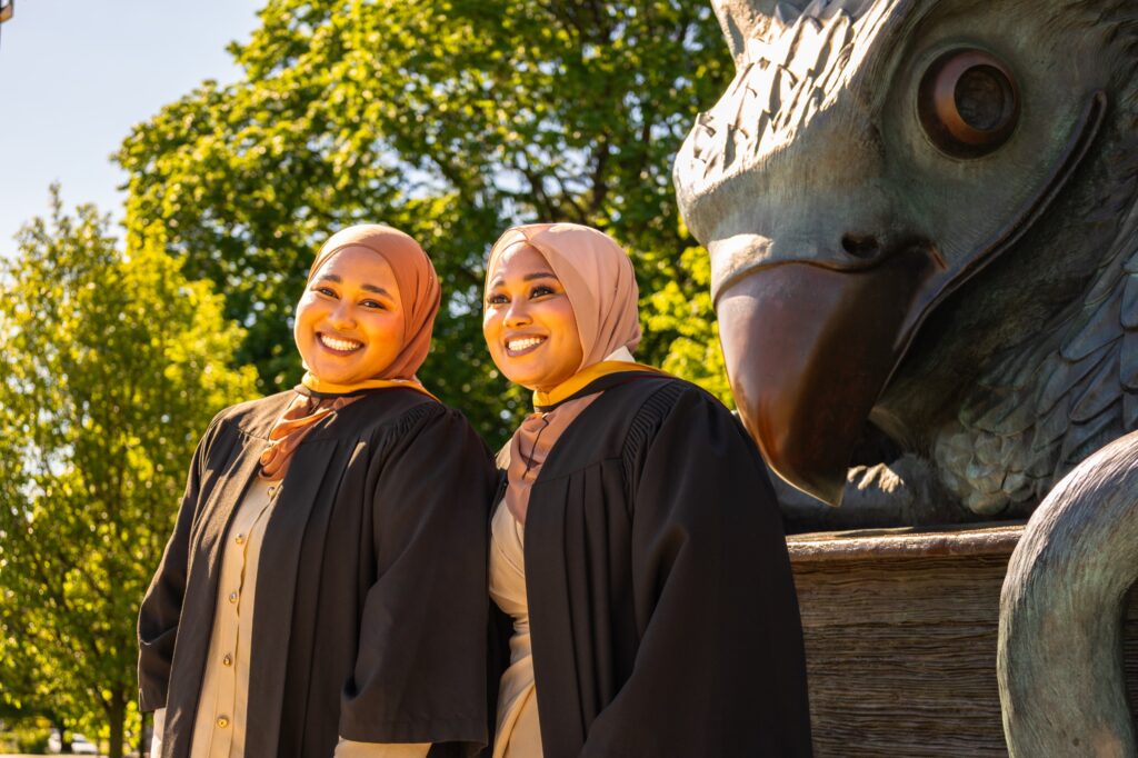 Two graduates wearing hijabs and black gowns smiling beside the bronze gryphon statue at the University of Guelph.