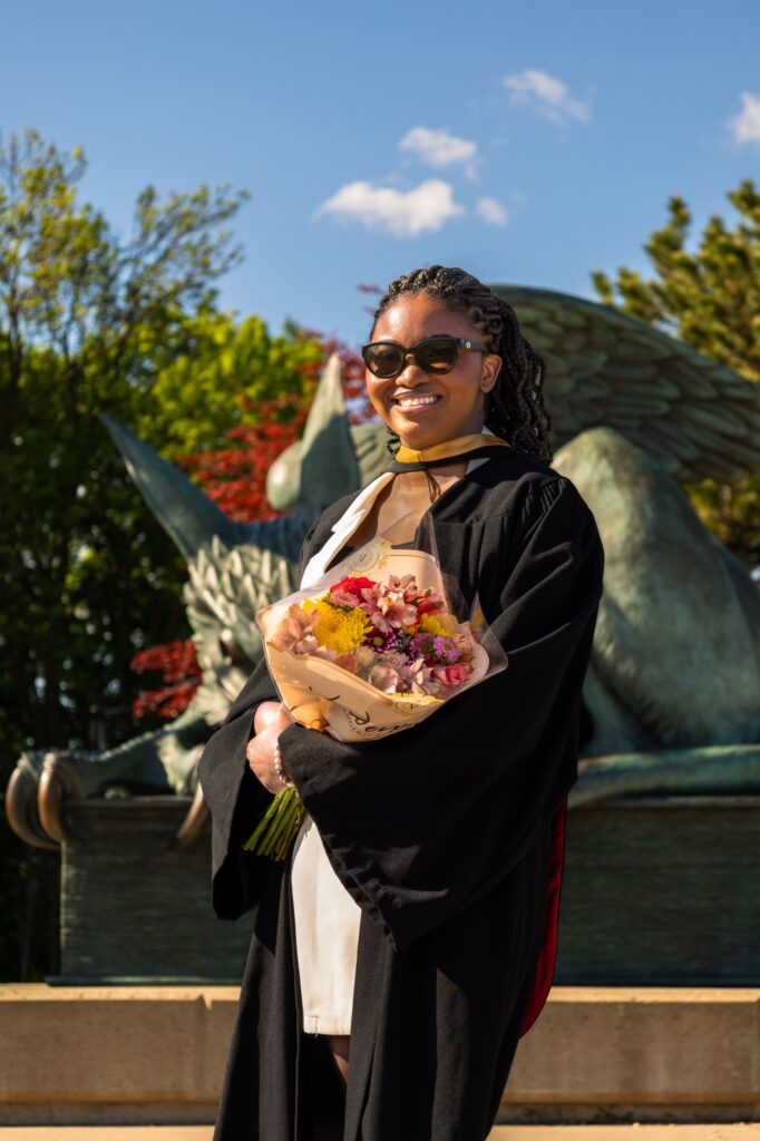A graduate in a black gown and sunglasses holding a bouquet of flowers in front of the bronze gryphon statue at the University of Guelph.
