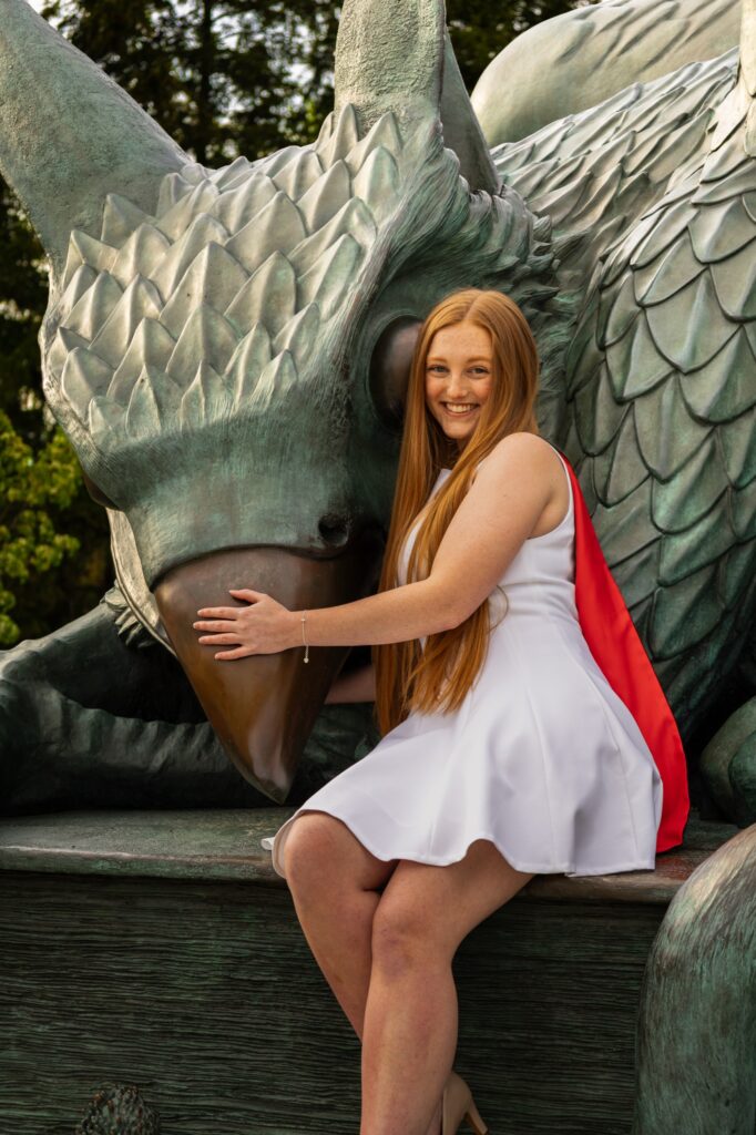 A graduate with long red hair in a white dress with a red stole sitting on the base of the bronze gryphon statue at the University of Guelph.