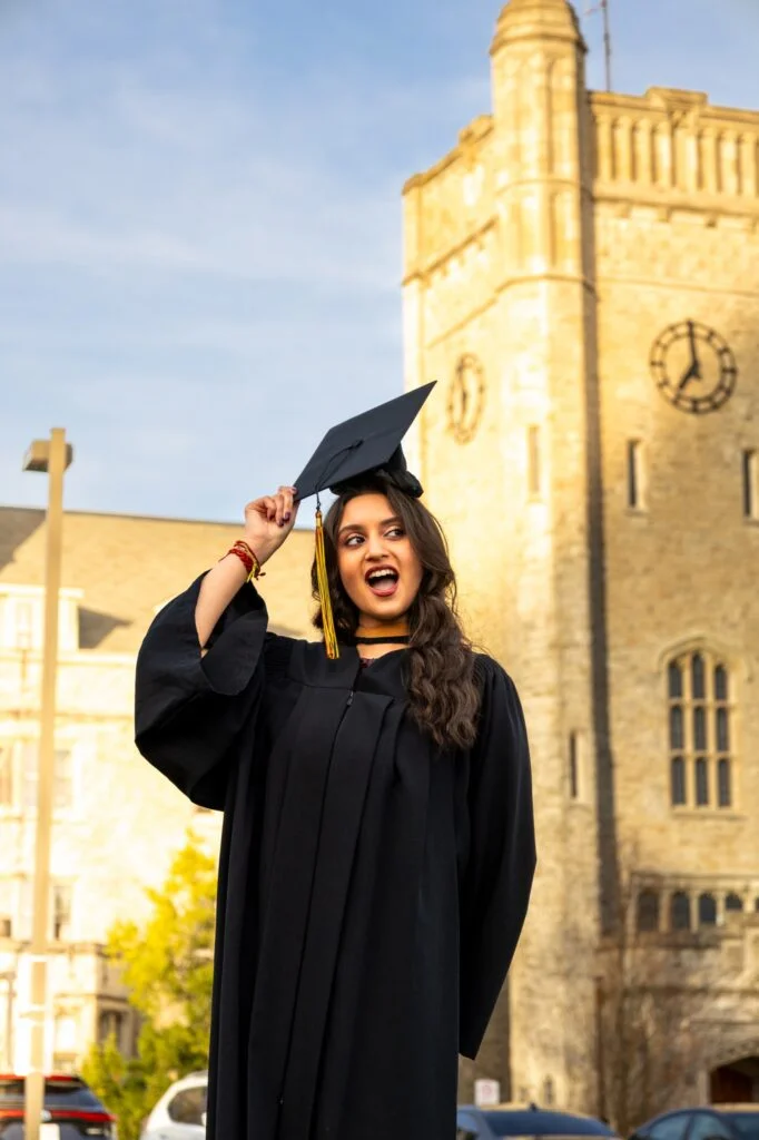 A graduate in a black gown holding her cap on her head in front of the Johnston Hall clock tower at the University of Guelph.