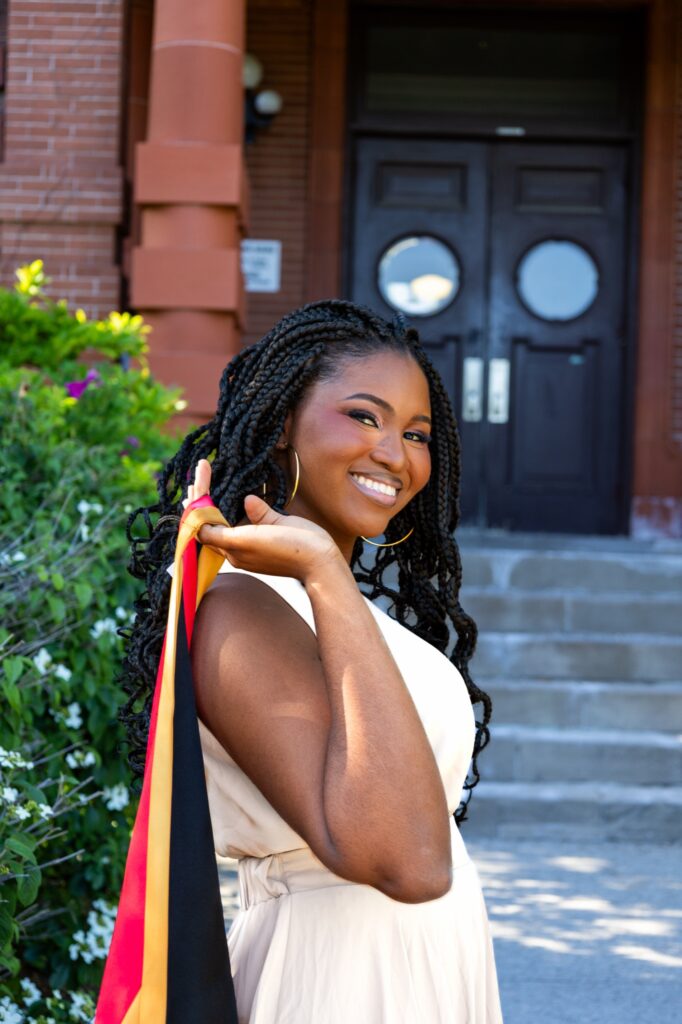 A smiling graduate in a cream dress holding a red, gold and black stole over her shoulder in front of a brick building entrance.