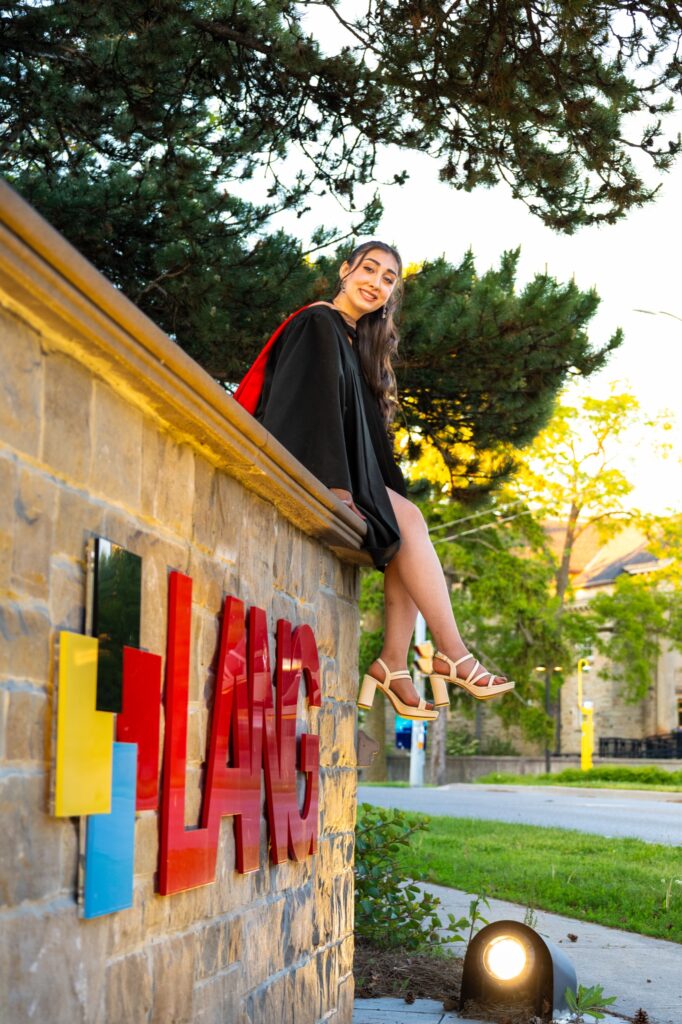 A graduate in a black gown with a red stole sitting on the stone wall of the Lang School of Business sign at the University of Guelph.