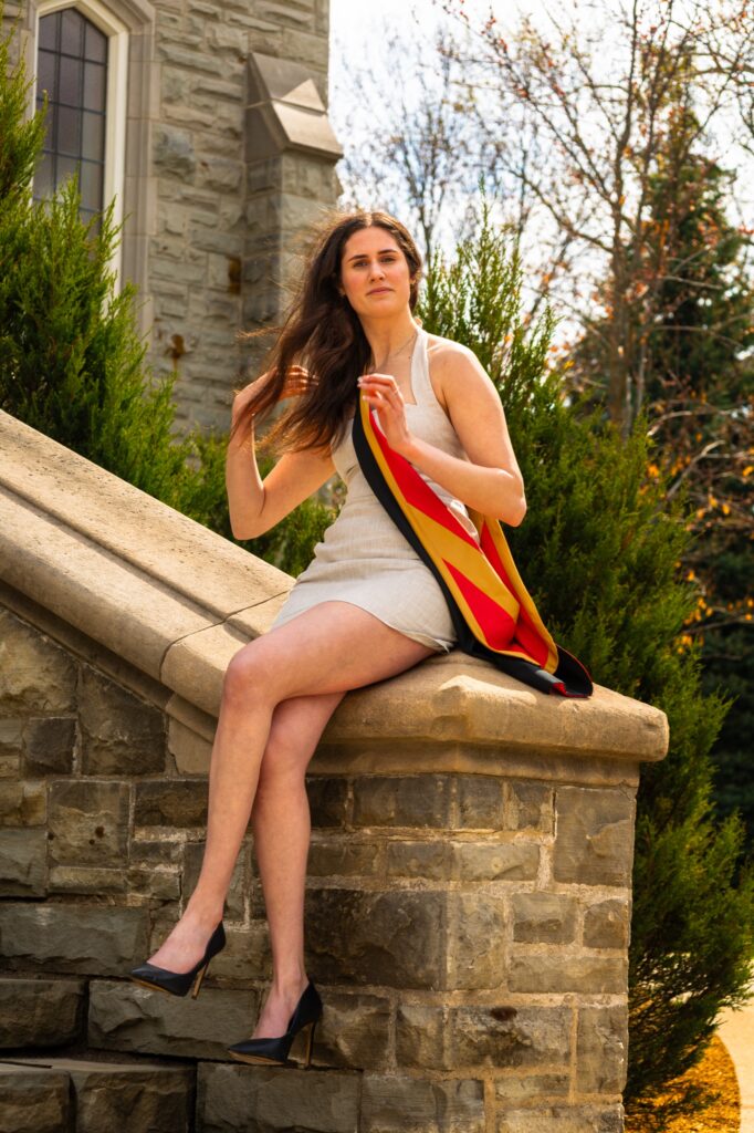 A graduate in a beige dress seated on the stone balustrade of a limestone building with a red, gold and black stole beside her.