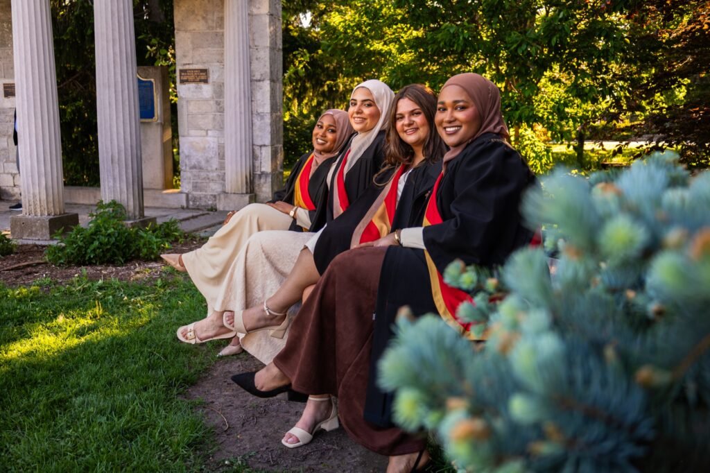 Four graduates in black gowns with red and gold hoods, two wearing hijabs, seated together at the Portico columns at the University of Guelph.