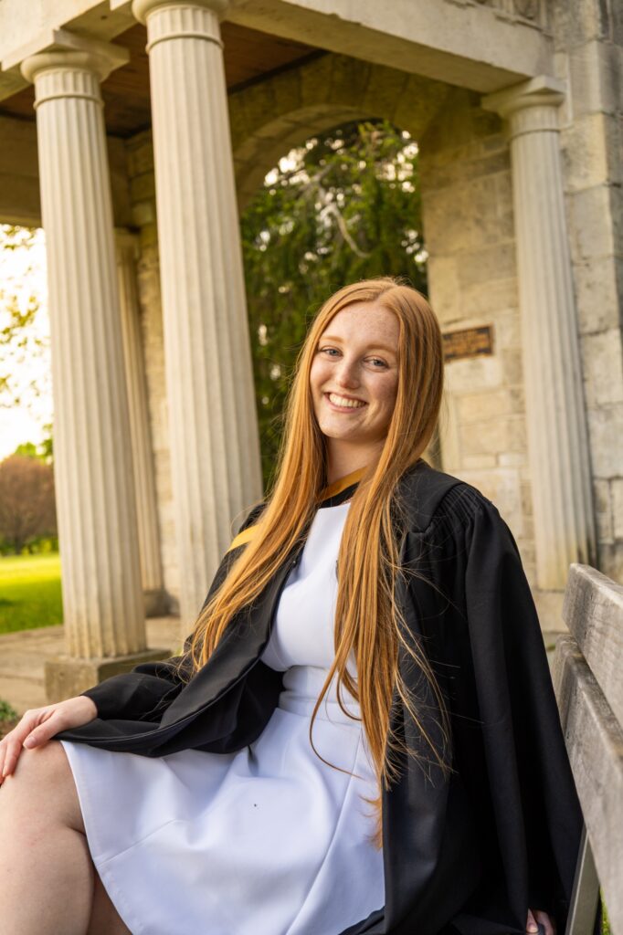 A graduate with long red hair in a white dress and black gown with a gold hood seated on a bench in front of the Portico columns at the University of Guelph.