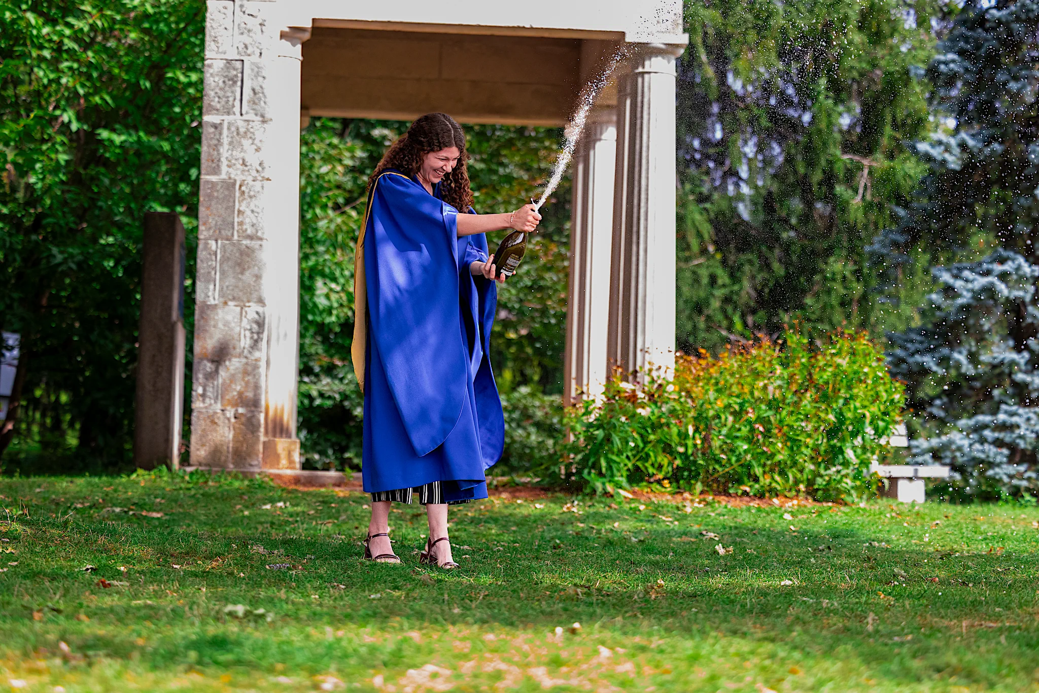 A University of Guelph graduate in regalia opening a bottle of champagne by a stone gateway on campus.