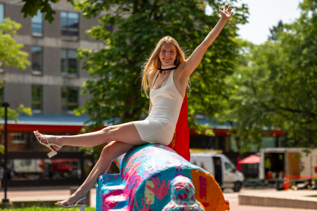 A smiling graduate in a white dress and red stole perched on the colourful painted Cannon with one arm stretched up