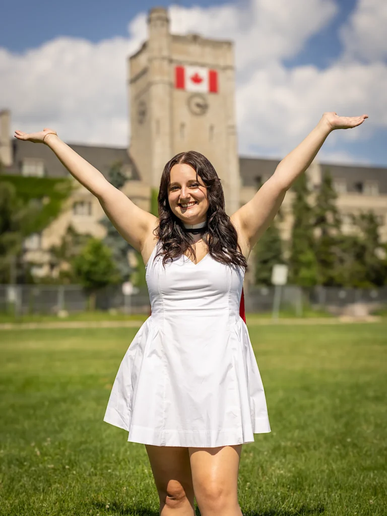 A University of Guelph graduate in regalia with her arms wide on the green in front of Johnston Hall.