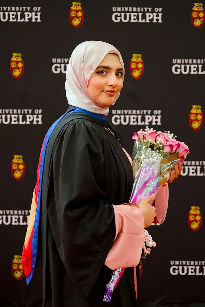 A University of Guelph graduate in regalia with a bouquet at the convocation backdrop.