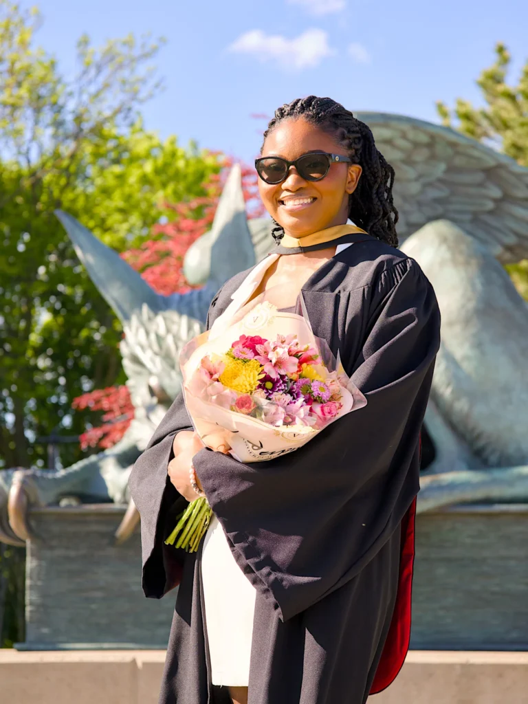 A University of Guelph graduate in regalia holding a bouquet in front of the Gryphon statue.