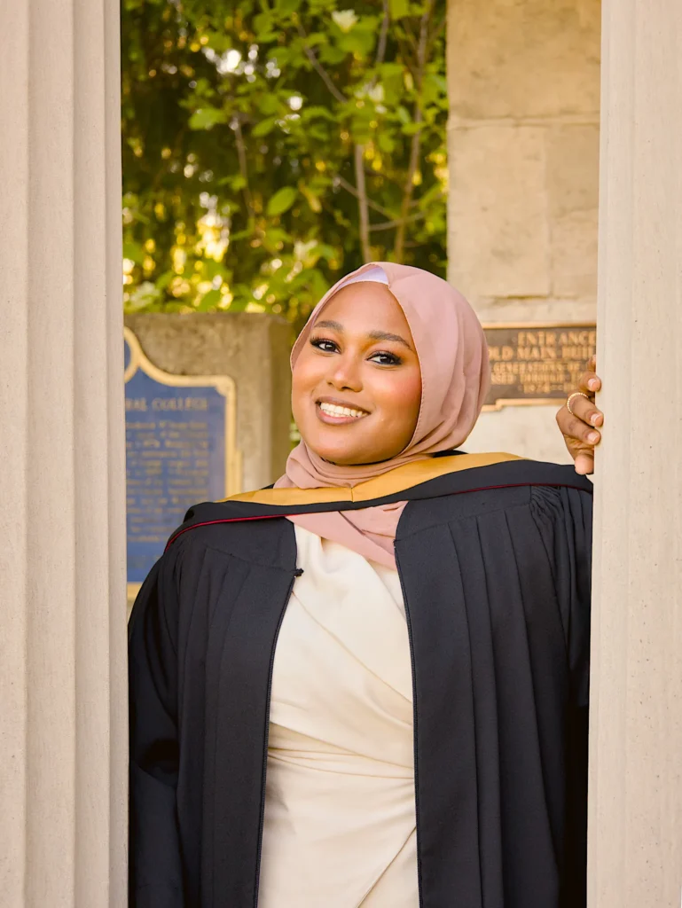 A University of Guelph graduate in regalia leaning on a column of the stone colonnade.