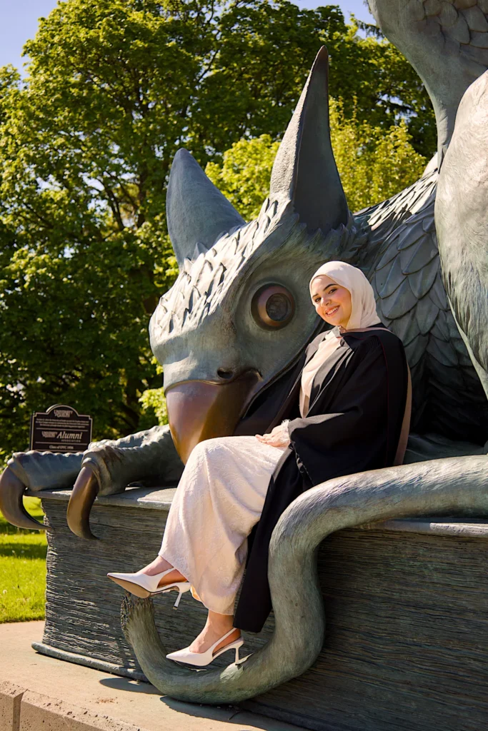 A University of Guelph graduate in regalia sitting in the lap of the Gryphon statue.