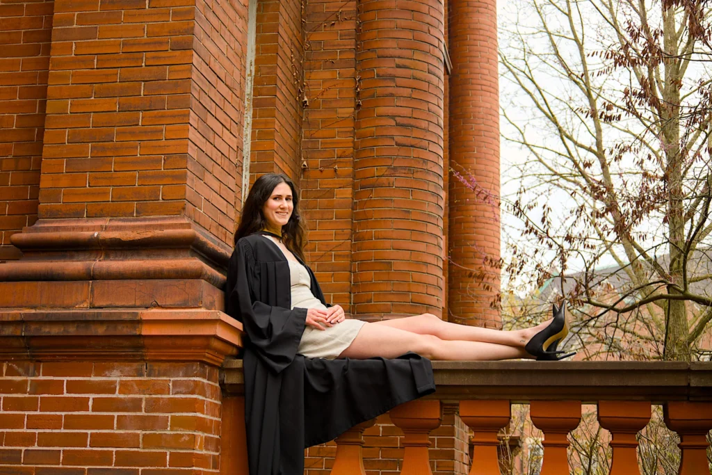 A University of Guelph graduate in regalia lying back along the balustrade of the red-brick hall.
