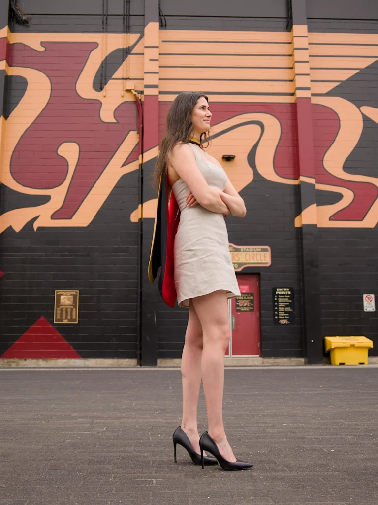 A University of Guelph graduate in regalia with her arms crossed in front of the campus mural wall.