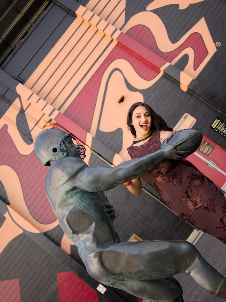 A University of Guelph graduate in regalia beside a bronze sculpture against the campus mural wall.
