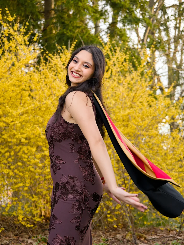 A University of Guelph graduate with her regalia catching the wind beside a forsythia in full bloom.